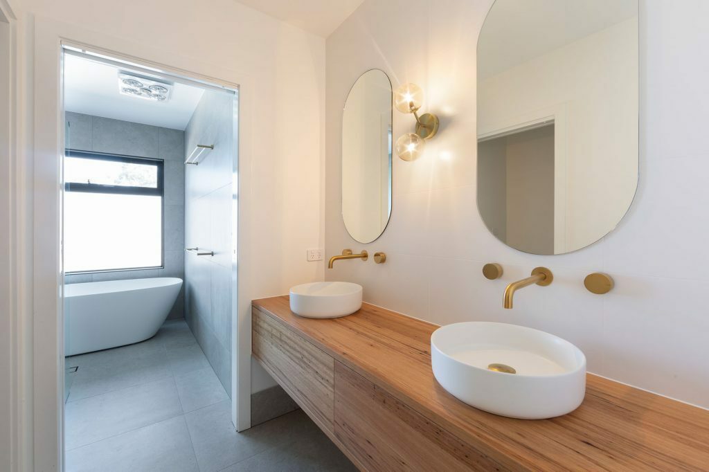 Homes by Howe, knock down rebuild, Duffy, bathroom, slate tiling on floor looking towards white freestanding bath, golden taps, oval mirrors above each sink, slate tiling on floor